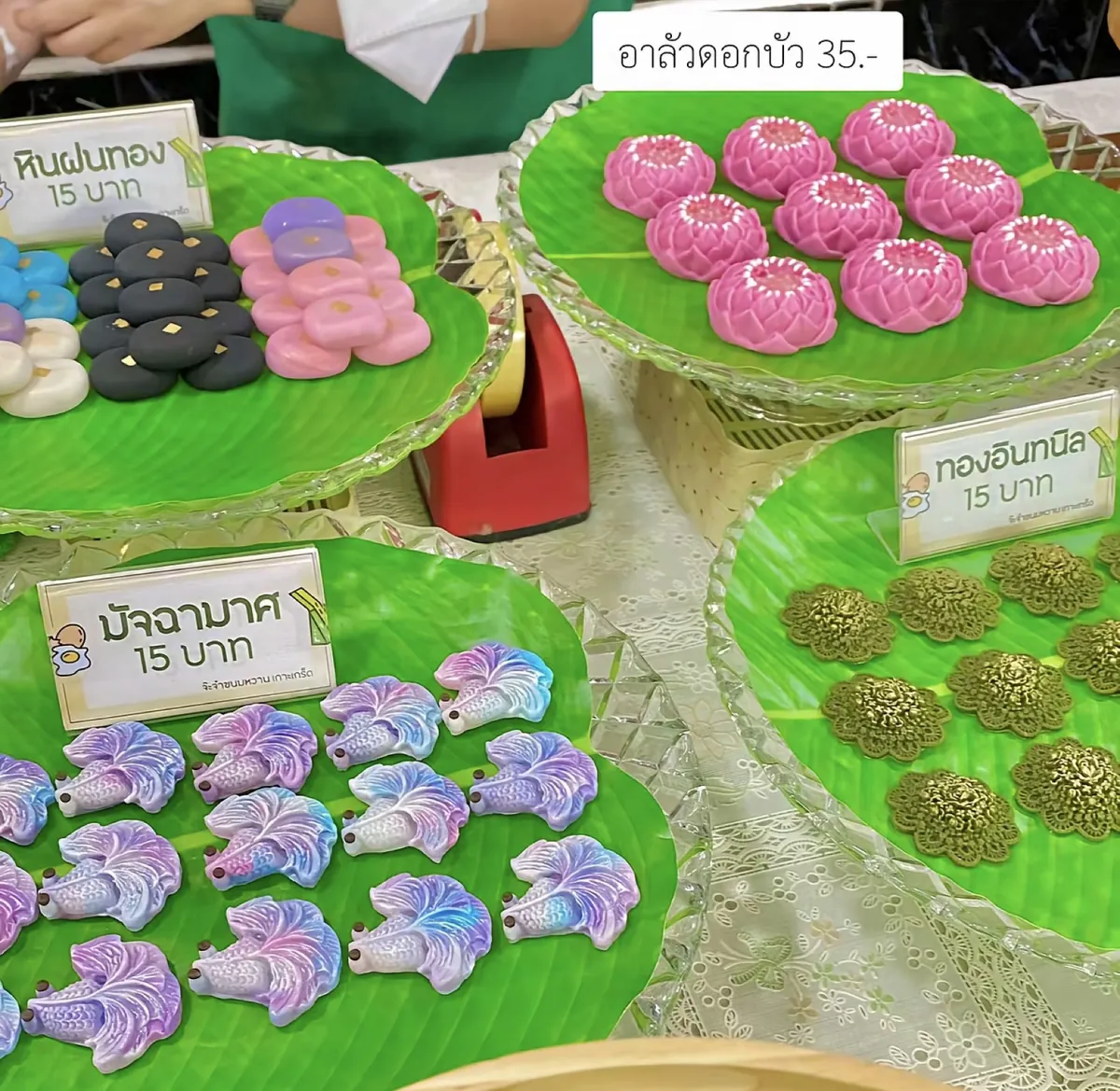 ถาดขนมวางบนใบตองสี่ถาด มีขนมรูปดอกบัวสีชมพู ขนมรูปปลากัดสีม่วงฟ้า ขนมดอกไม้สีทอง และขนมกลม ๆ หลายสี แต่ละถาดมีป้ายบอกชื่อและราคาเสียบไว้