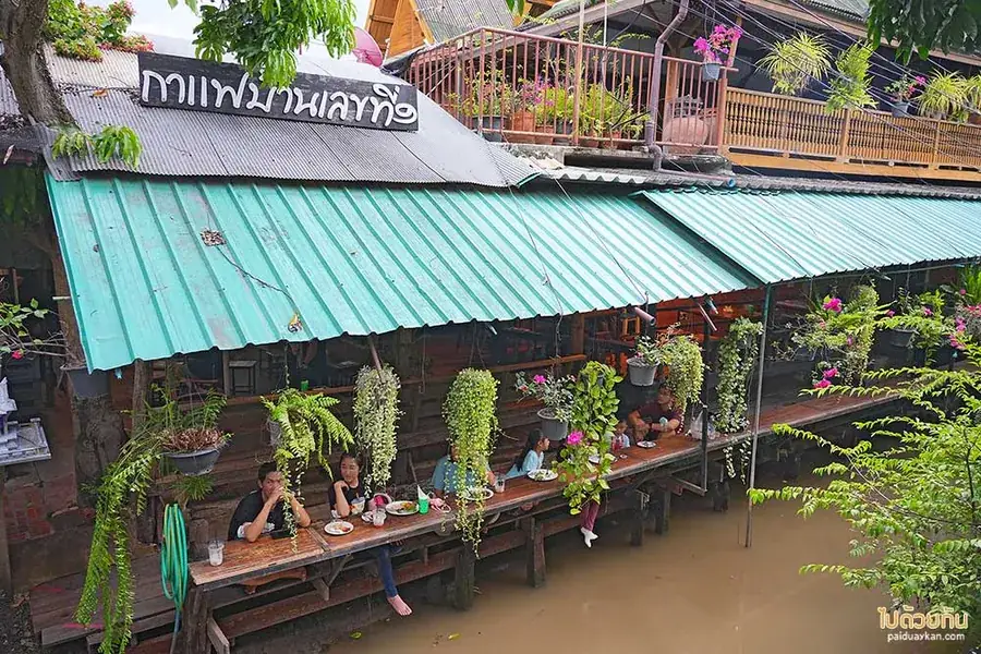 ร้านกาแฟไม้หลังคาสังกะสีสีเขียวมิ้นต์ ยื่นระเบียงออกไปเหนือคลอง มีคนนั่งกินอาหารเรียงกันตามโต๊ะยาว และมีกระถางไม้เลื้อยแขวนตลอดแนว