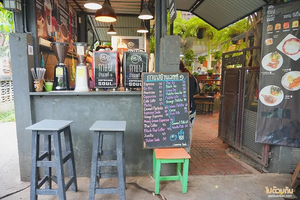 หน้าร้านกาแฟเปิดโล่ง มีเครื่องบดกาแฟวางบนเคาน์เตอร์ปูน เก้าอี้สูงสองตัว และกระดานดำเขียนรายการเครื่องดื่มพร้อมราคาตั้งอยู่ข้างทางเดิน