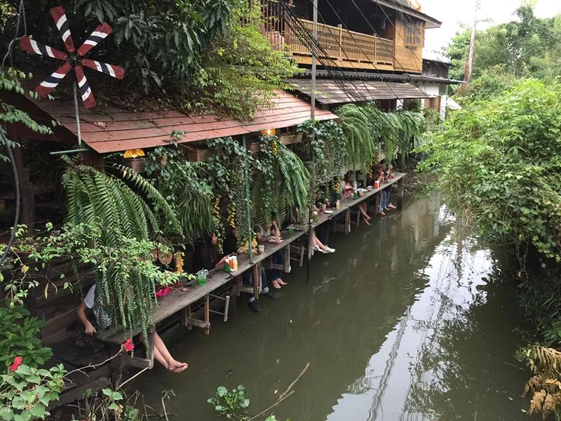แถวที่นั่งริมคลองใต้ชายคาไม้ คนนั่งเรียงกันหันหน้าออกทางน้ำ ห้อยขาลงไปเหนือผิวน้ำ มีเฟินและไม้เลื้อยแขวนบังแดดอยู่ด้านบน