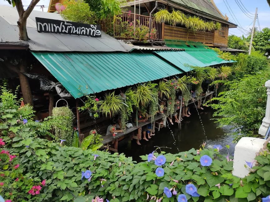 ร้านไม้หลังคาสังกะสีสีเขียว มีป้ายชื่อร้านอยู่บนหลังคา แถวที่นั่งยื่นออกไปเหนือคลอง คนนั่งห้อยขาเรียงกัน มีกระถางไม้เลื้อยแขวนตลอดแนวและดอกไม้สีม่วงอยู่หน้าภาพ
