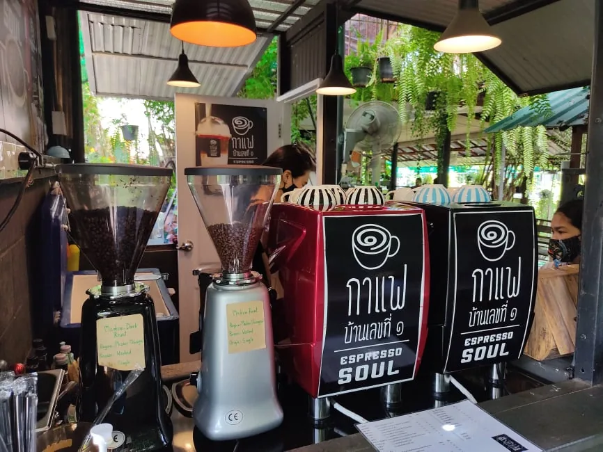 เคาน์เตอร์กาแฟ มีเครื่องบดเมล็ดสองเครื่องวางเรียง ป้ายชื่อร้านสีดำเขียนว่า กาแฟบ้านเลขที่ ๑ ESPRESSO SOUL และพนักงานยืนทำเครื่องดื่มอยู่ด้านหลัง