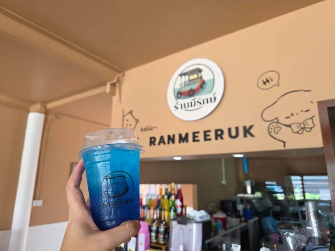 มือถือแก้วเครื่องดื่มสีฟ้าที่พิมพ์โลโก้ร้านมีรักษ์ ด้านหลังเป็นผนังสีครีมติดป้ายชื่อร้านทั้งภาษาไทยและคำว่า RANMEERUK พร้อมเคาน์เตอร์เครื่องดื่ม