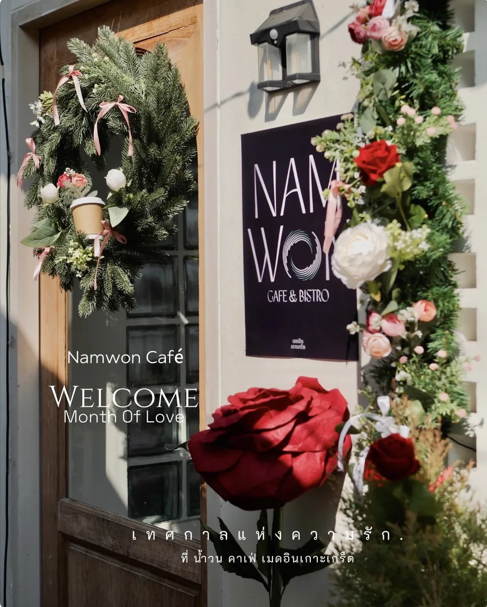 ประตูไม้หน้าร้าน มีป้ายสีดำเขียนว่า NAMWON CAFE & BISTRO ติดอยู่บนผนัง ประดับด้วยพวงมาลัยใบสนและดอกกุหลาบสีแดงกับชมพูรอบกรอบประตู