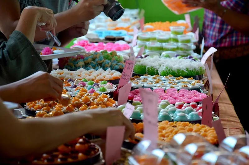 โต๊ะขายขนมไทยหลายสิบถาด มีขนมสีชมพู เขียว ส้ม และขาว วางเรียงเป็นแถวยาว มีคนยืนเลือกซื้อและยกกล้องถ่ายรูปอยู่ด้านหลัง