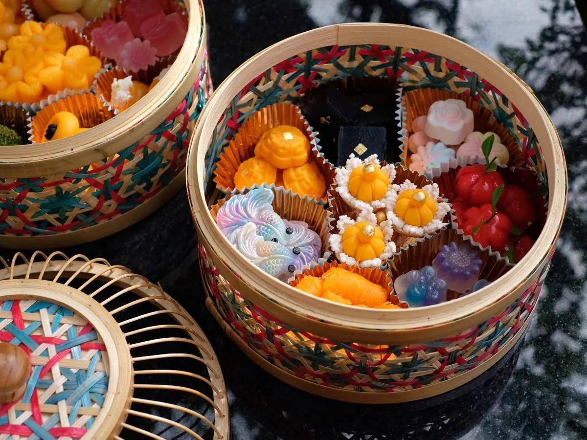 ขนมไทยหลายชนิดจัดใส่กระทงกระดาษเรียงในกระติบไม้ไผ่สานลายสี มีทั้งขนมสีเหลืองทอง ขนมรูปดอกไม้สีชมพู ขนมลูกกลมสีแดง และขนมสีดำปิดทองคำเปลว