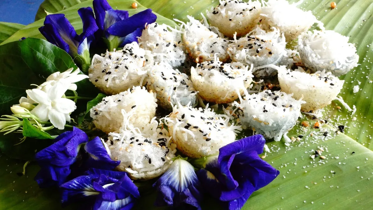 ขนมครกหลายชิ้นวางเรียงบนใบตอง แต่ละชิ้นโรยมะพร้าวขูดกับงาดำและน้ำตาล ประดับรอบด้วยดอกอัญชันสีม่วงและดอกมะลิสีขาว