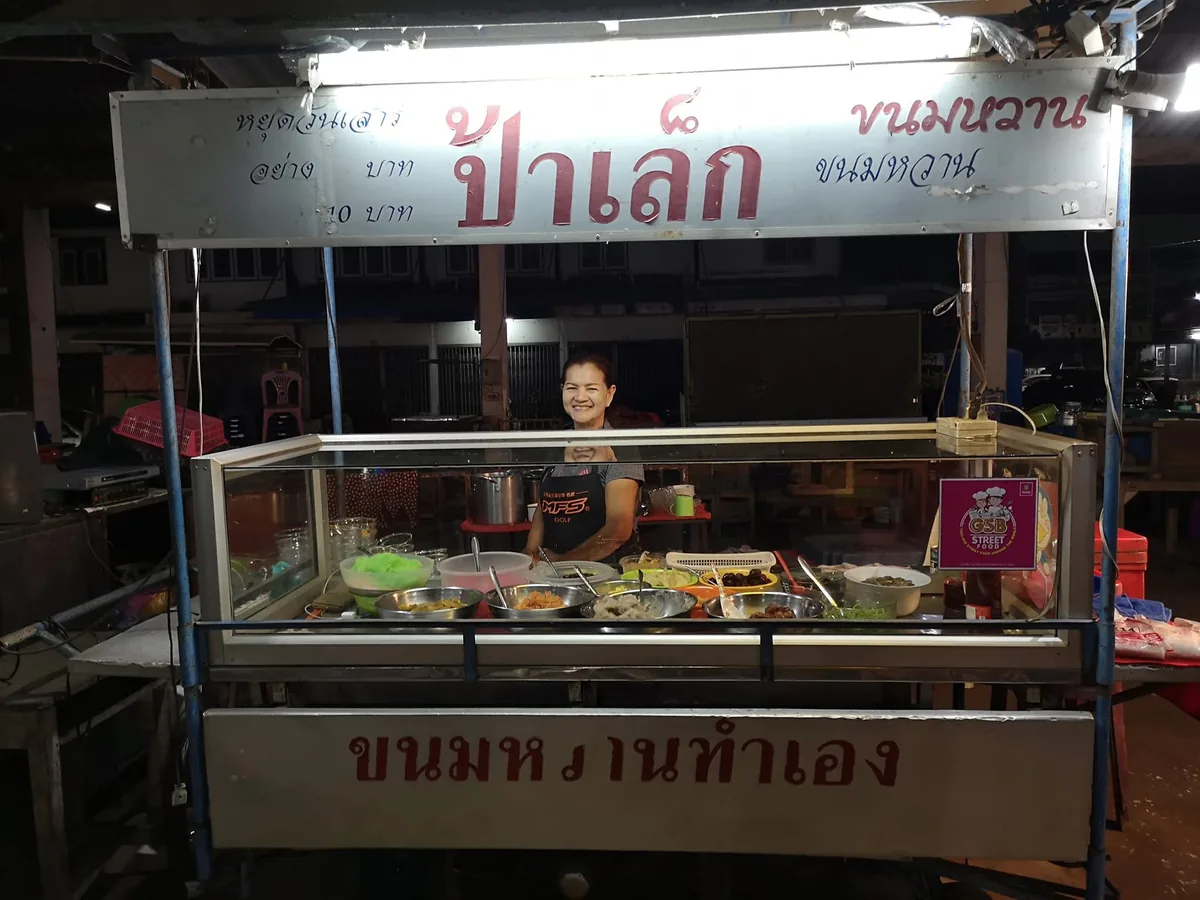 รถเข็นขนมหวานตอนกลางคืน มีป้ายไฟเขียนว่า ป้าเล็ก ขนมหวาน อยู่ด้านบน และป้ายคำว่า ขนมหวานทำเอง อยู่ด้านล่าง ในตู้กระจกมีถ้วยเครื่องขนมวางเรียงหลายอย่าง