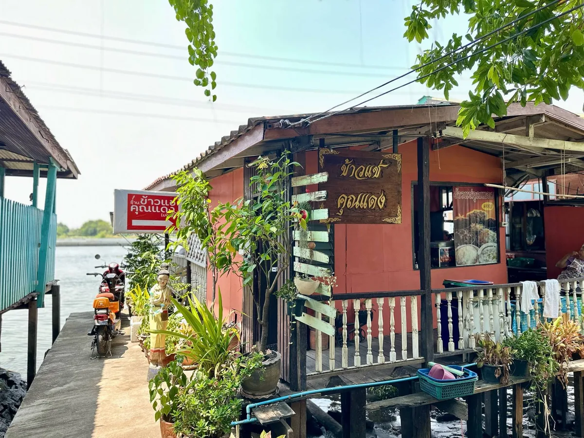 ร้านไม้ริมน้ำทาสีชมพูอมส้ม มีป้ายไม้แกะสลักเขียนว่า ข้าวแช่คุณแดง ติดอยู่หน้าร้าน ระเบียงลูกกรงสีขาวยื่นออกไปเหนือน้ำ และมีกระถางต้นไม้วางเรียงตามทางเดินปูน