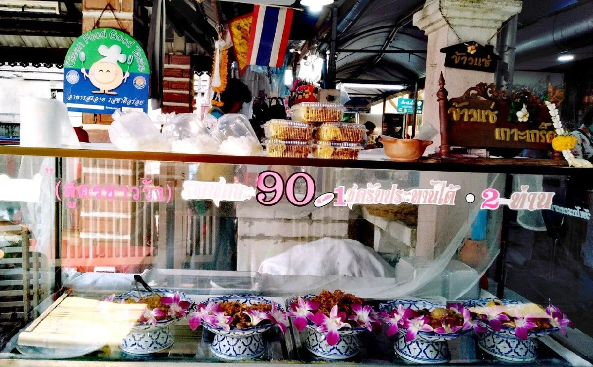 ตู้กระจกหน้าร้าน ด้านในเรียงถ้วยลายครามใส่เครื่องเคียงประดับดอกกล้วยไม้สีม่วง มีป้ายราคาติดกระจกเขียนว่าสูตรชาววัง 90 บาท และป้ายไม้แกะสลักคำว่าข้าวแช่ เกาะเกร็ด อยู่ด้านบน