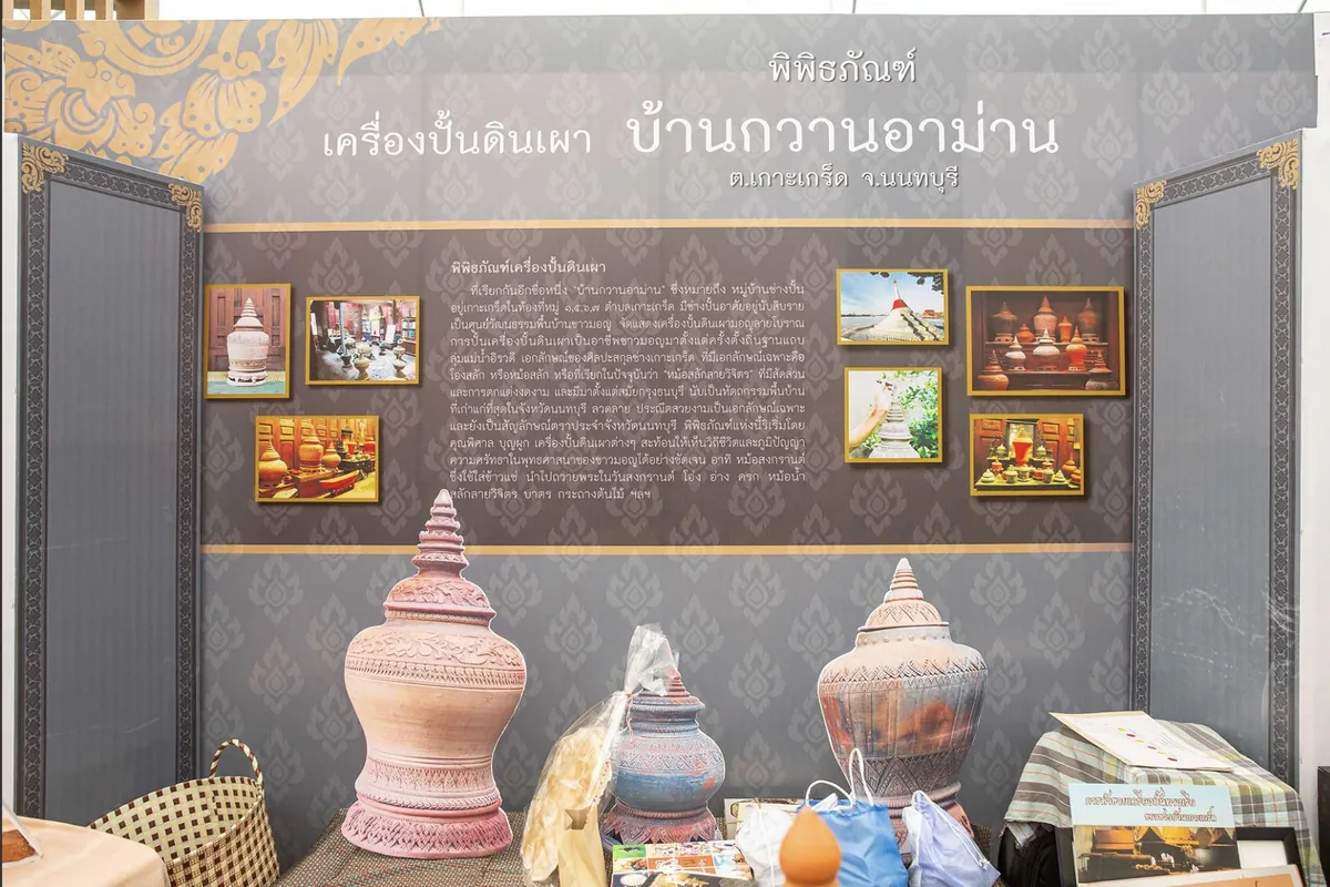 บูธพิพิธภัณฑ์เครื่องปั้นดินเผาบ้านกวานอาม่าน ตำบลเกาะเกร็ด จังหวัดนนทบุรี มีป้ายชื่อและภาพประกอบติดผนัง ด้านหน้าตั้งโอ่งดินเผาสลักลายสามใบ
