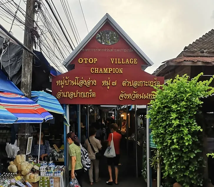 ซุ้มทางเข้าตลาด ป้ายไม้สีแดงเขียนว่า OTOP Village Champion หมู่บ้านโอ่งอ่าง หมู่ ๗ ตำบลเกาะเกร็ด อำเภอปากเกร็ด จังหวัดนนทบุรี ด้านหลังเป็นทางเดินแคบที่มีคนเดินอยู่