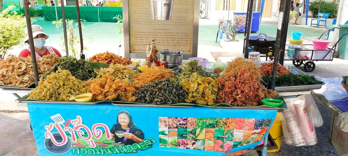 แผงของทอดที่ตั้งอยู่ในลานวัด มีถาดผักทอดและดอกไม้ทอดหลายกองเรียงยาวเต็มแผง ด้านหน้าติดป้ายไวนิลสีฟ้าเขียนว่าป้าอู๊ด ทอดมันหน่อกะลา พร้อมรูปเจ้าของร้านและตารางรูปเมนู