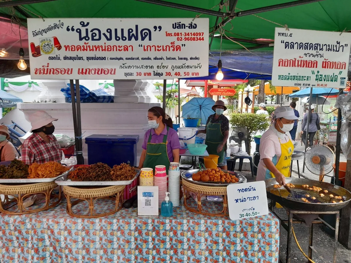 แผงขายใต้เต็นท์ มีป้ายชื่อร้านน้องแฝดทอดมันหน่อกะลาเกาะเกร็ดแขวนอยู่ด้านบน ระบุราคาชุดละ 30 บาท และใส่ถุง 30 40 50 บาท ด้านล่างเป็นถาดของทอดหลายอย่างกับกระทะทอดที่กำลังทอดอยู่