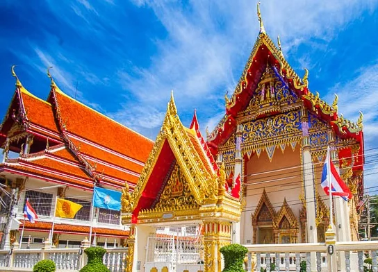 อาคารทรงไทยของวัด หลังคาซ้อนชั้นมุงกระเบื้องสีส้มสลับแดง หน้าบันปิดทองประดับกระจกสี มีซุ้มประตูยอดแหลมปิดทองอยู่ตรงกลาง ด้านหน้าปักธงชาติไทยและธงสีเหลืองเรียงเป็นแถว ท้องฟ้าสีฟ้าจัด