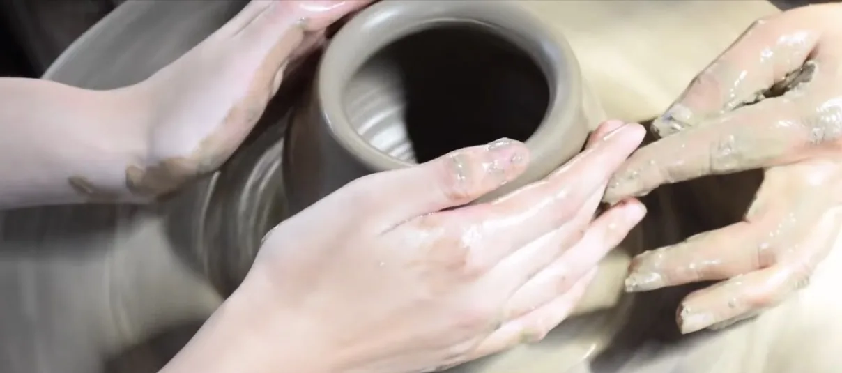 สองมือประคองดินบนแป้นหมุน ดินขึ้นรูปเป็นทรงกระบอกปากกว้าง มือเปื้อนดินเหนียวสีเทาทั้งสองข้าง ขอบแป้นที่หมุนอยู่เบลอเป็นวง